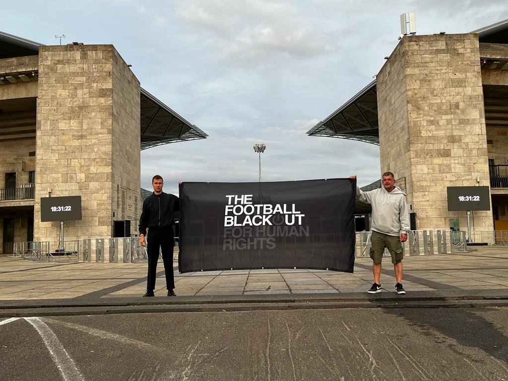 Zwei Männer halten in Banner mit dem Schriftzug "The Football Blackout for Human Rights" hoch, wahrscheinlich vor dem Olympiastadion in Berlin.