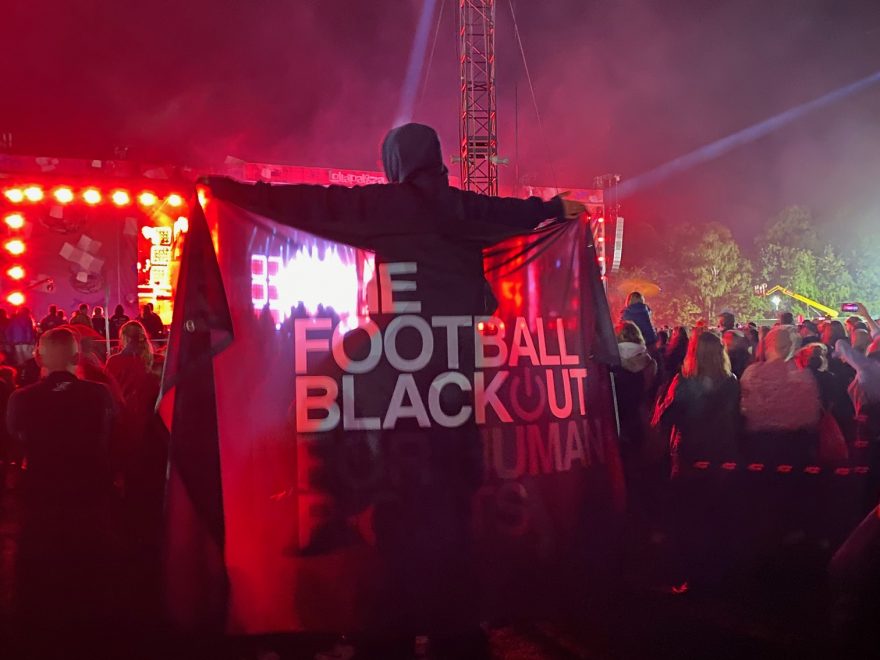 Nachts cor einer Bühne mit Publikum, ein Mensch hält das Banner mit dem Schriftzug "The Football Blackout for Human Rights" hoch.