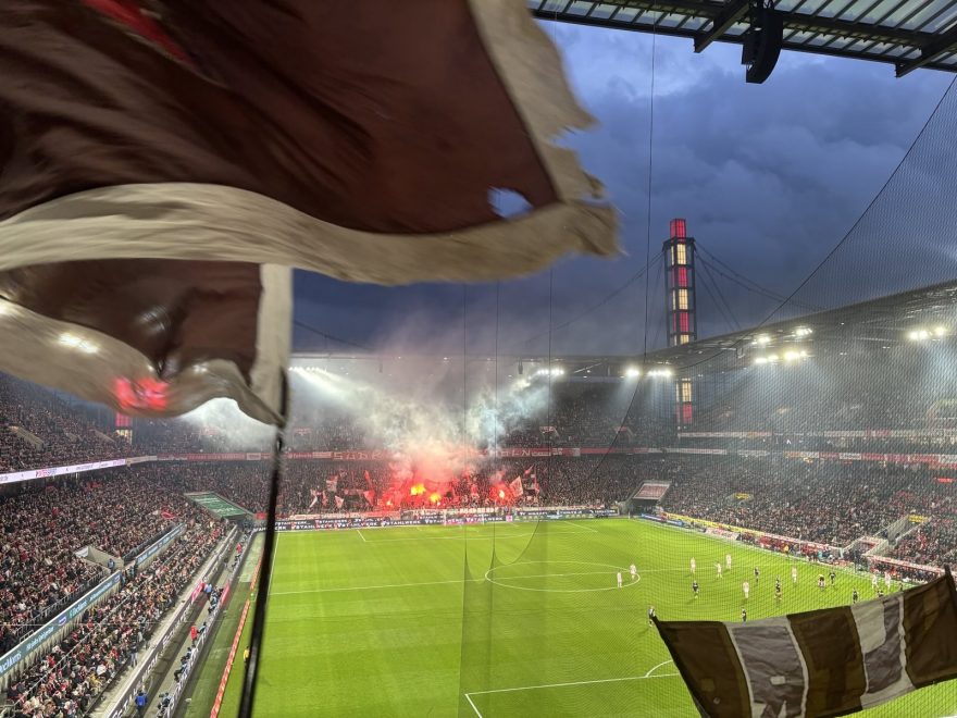 Im Vordergrund wehende Fahnen im St. Pauli Block, im Hintergrund Pyro im Kölner Block im Rhein Energie Stadion.