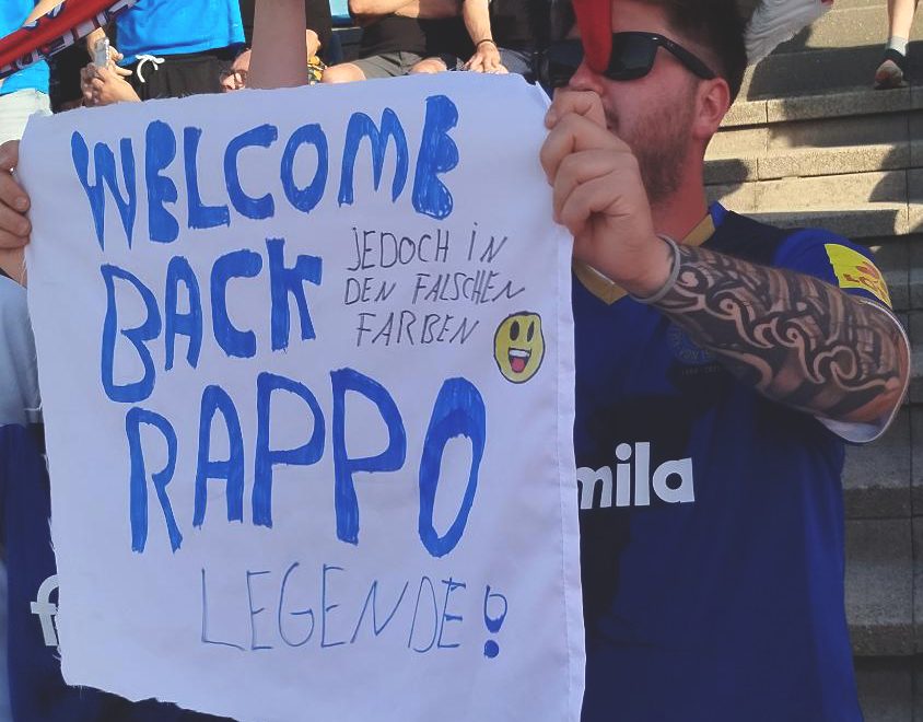 Ein Kieler Fan mit Storch-Hut hält ein großes Papier in den Händen auf dem steht: "Welcome back Rappo Legende" und in klein "jedoch in den falschen Farben".